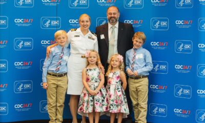 A mom in white uniform standing with her husband and two sets of twins (older boys and young girls) in front of a blue background.