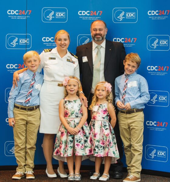 A mom in white uniform standing with her husband and two sets of twins (older boys and young girls) in front of a blue background.