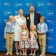 A mom in white uniform standing with her husband and two sets of twins (older boys and young girls) in front of a blue background.