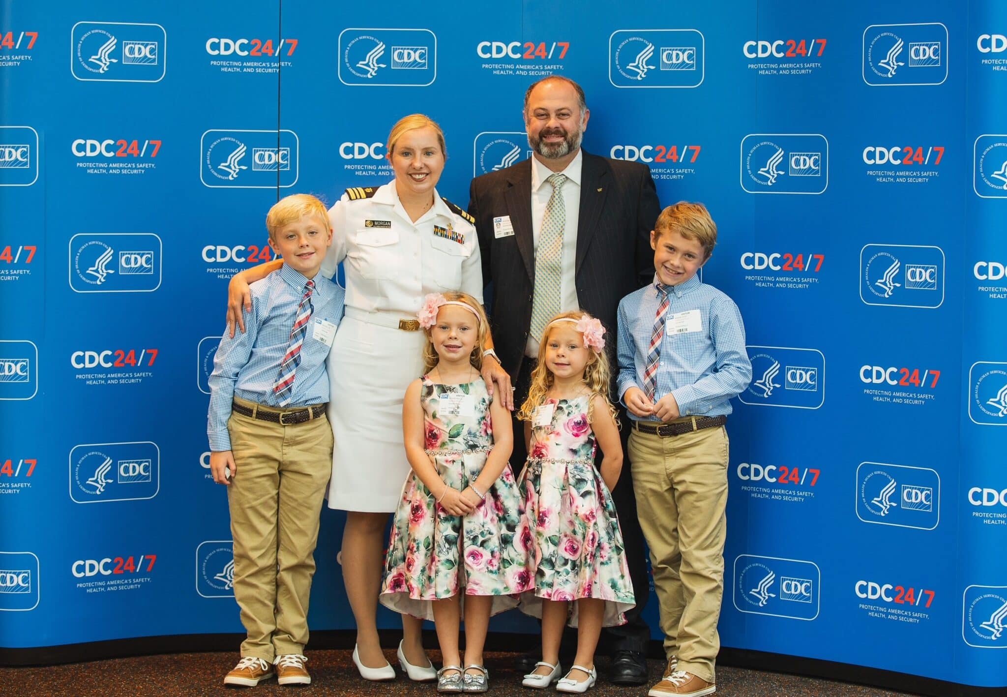 A mom in white uniform standing with her husband and two sets of twins (older boys and young girls) in front of a blue background.