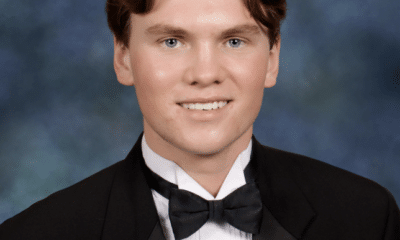 Senior portrait of a young white man wearing a black suit, white button up shirt and black bowtie. He has short brown hair parted in the middle, and he's smiling as he sits in front of a muted blue background.