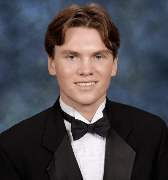 Senior portrait of a young white man wearing a black suit, white button up shirt and black bowtie. He has short brown hair parted in the middle, and he's smiling as he sits in front of a muted blue background.