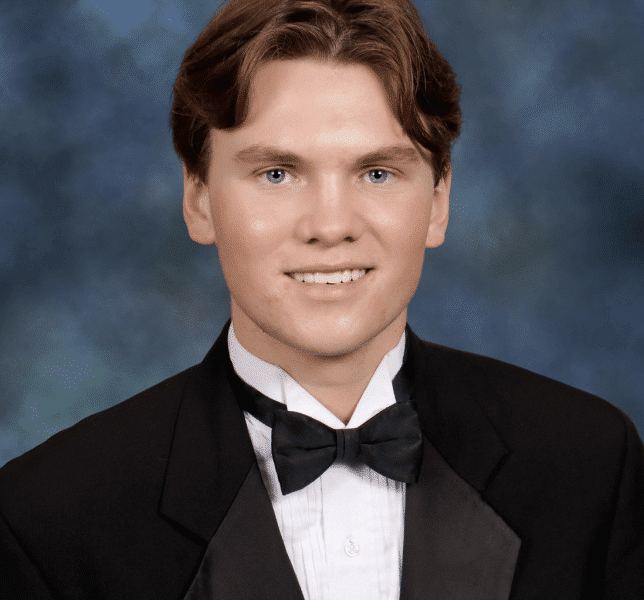 Senior portrait of a young white man wearing a black suit, white button up shirt and black bowtie. He has short brown hair parted in the middle, and he's smiling as he sits in front of a muted blue background.