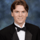 Senior portrait of a young white man wearing a black suit, white button up shirt and black bowtie. He has short brown hair parted in the middle, and he's smiling as he sits in front of a muted blue background.