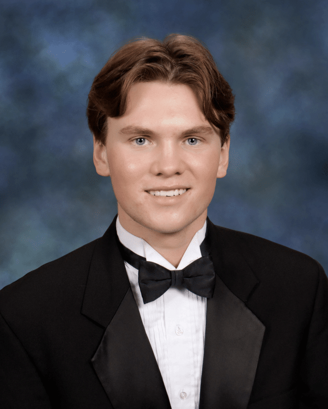 Senior portrait of a young white man wearing a black suit, white button up shirt and black bowtie. He has short brown hair parted in the middle, and he's smiling as he sits in front of a muted blue background.
