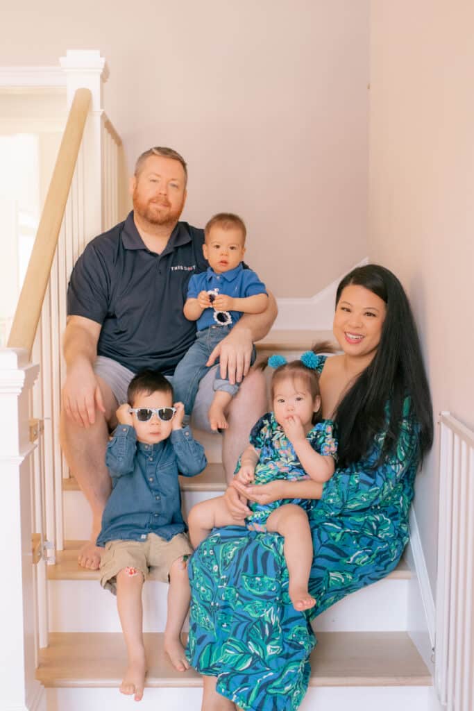 A family with mom, dad and three young kids. They're all dressed nicely for the photo and are sitting on the stairs of their home.