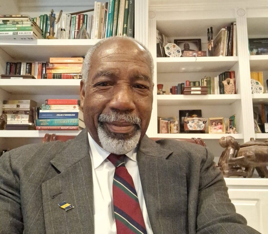 A distinguished Black man with short grey hair and grey facial hair. He's wearing a grey suite with white shirt, red and grey tie and a small yellow and blue lapel pin, sitting in front of white, built-in bookcases in his study.