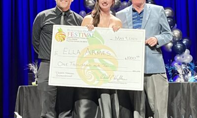 A young woman wearing a long, floor-length dress receiving a $1000 scholarship for drama. She's standing on stage with two men, holding a large version of the check. The background is a dark blue curtain.