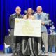 A young woman wearing a long, floor-length dress receiving a $1000 scholarship for drama. She's standing on stage with two men, holding a large version of the check. The background is a dark blue curtain.