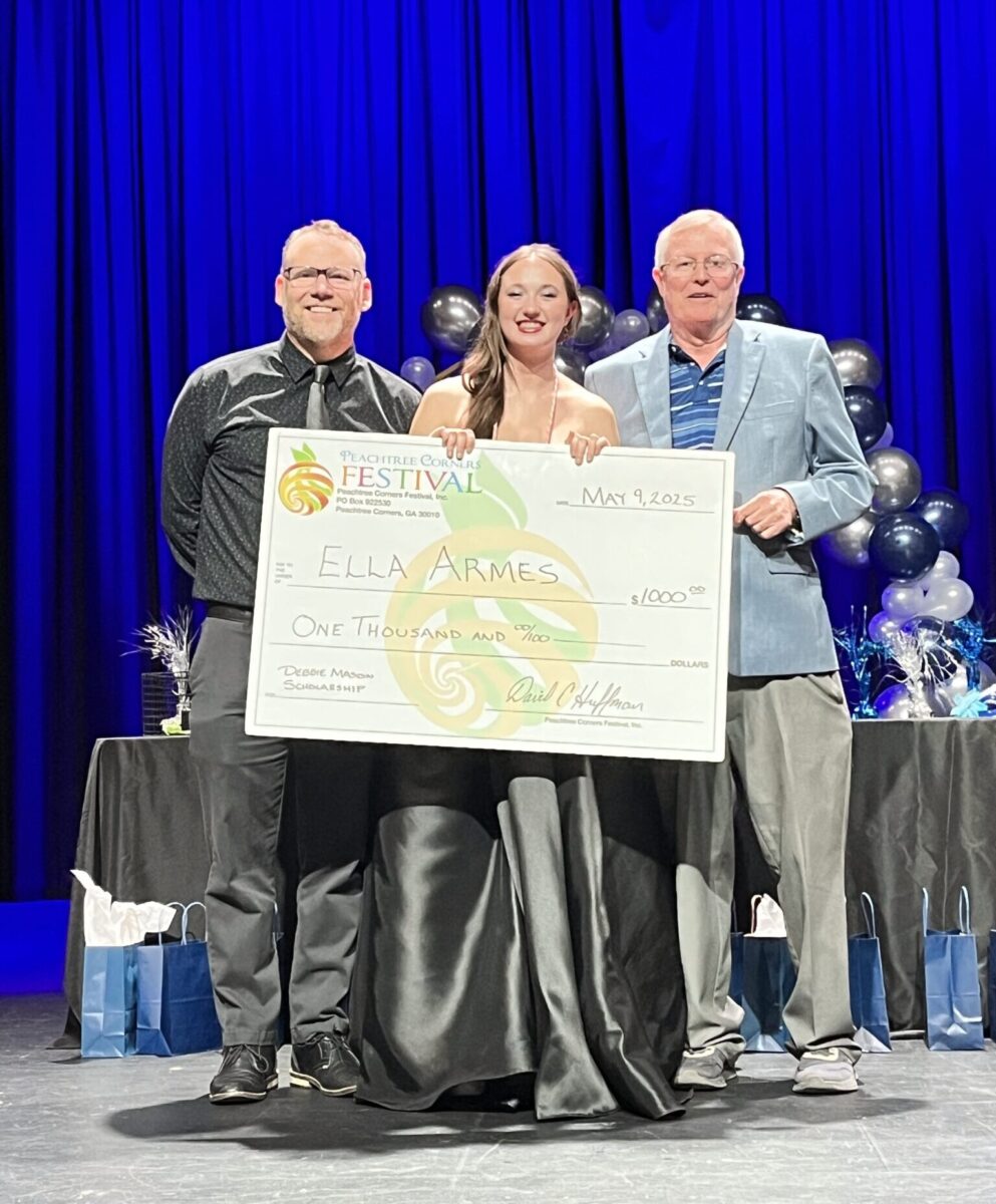 A young woman wearing a long, floor-length dress receiving a $1000 scholarship for drama. She's standing on stage with two men, holding a large version of the check. The background is a dark blue curtain.