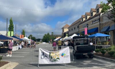 Outdoor farmer's market with tents lining the edges of a blocked off parking lot. There's signage, people walking about, and blue skies overhead.