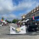 Outdoor farmer's market with tents lining the edges of a blocked off parking lot. There's signage, people walking about, and blue skies overhead.