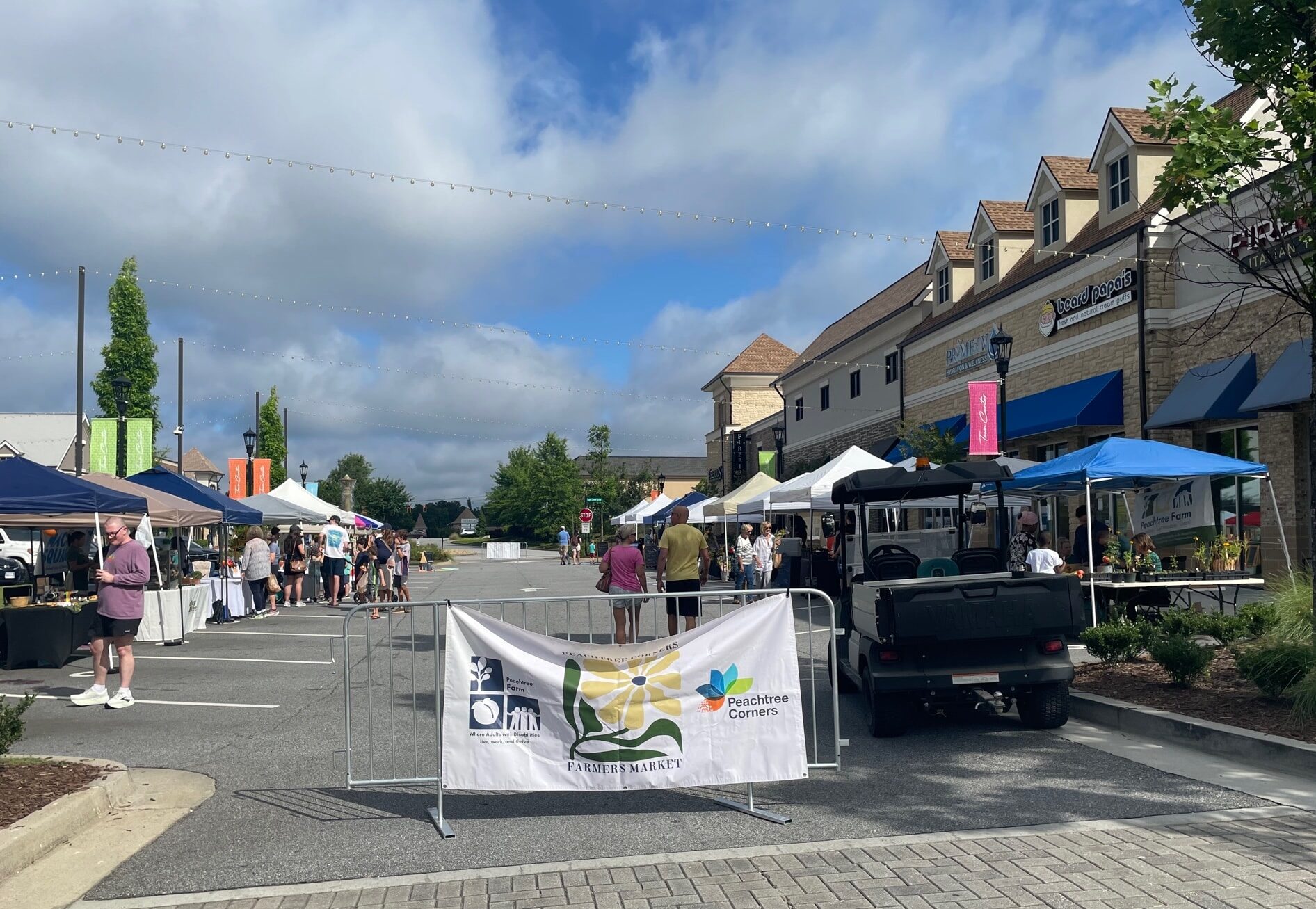 Outdoor farmer's market with tents lining the edges of a blocked off parking lot. There's signage, people walking about, and blue skies overhead.