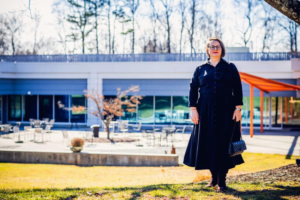 A middle aged woman with side-parted dark hair. She's wearing a dark colored dress and glasses, standing outdoors in the grass in front of Atlanta Tech Park in Peachtree Corners