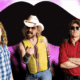 Mustache the Band standing in front of a glowing pink and white background with a large black mustache graphic.