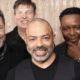 Four middle-aged men wearing black, standing together against a cream-colored backdrop and smiling at the camera.