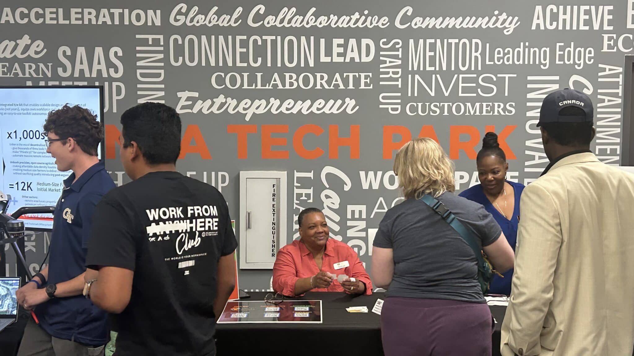 A person with short dark hair, wearing a red shirt sits at a table at a tech conference. Behind him there's a grey wall with words such as tech, connection, lead, collaborate and explore written in white. In front of him, several people are lined up to talk to him and others at the table.