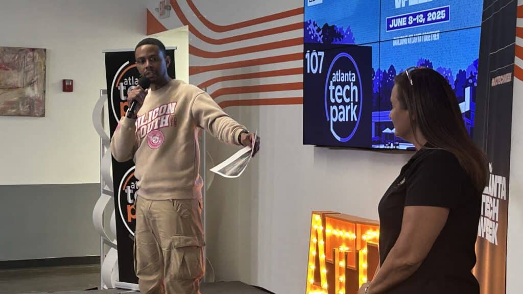 A young Black man wearing a tan colored outfit is holding a mic and speaking to a group of people at a tech conference. A woman with dark hair stands off to the side as he speaks.