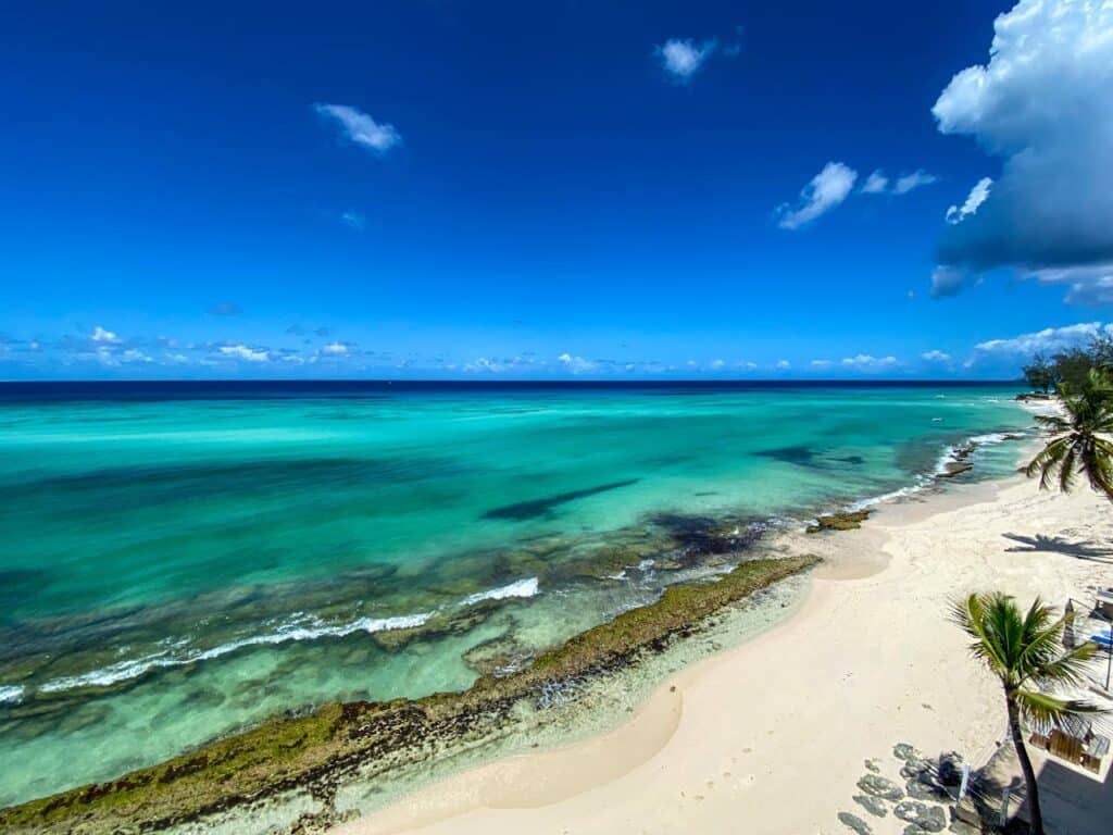 Aerial view of a stretch of beach in Barbados, with white sand, palm trees, a few lounger chairs and a wide expanse of turquise water. The sky overhead is deep blue with a few white clouds.