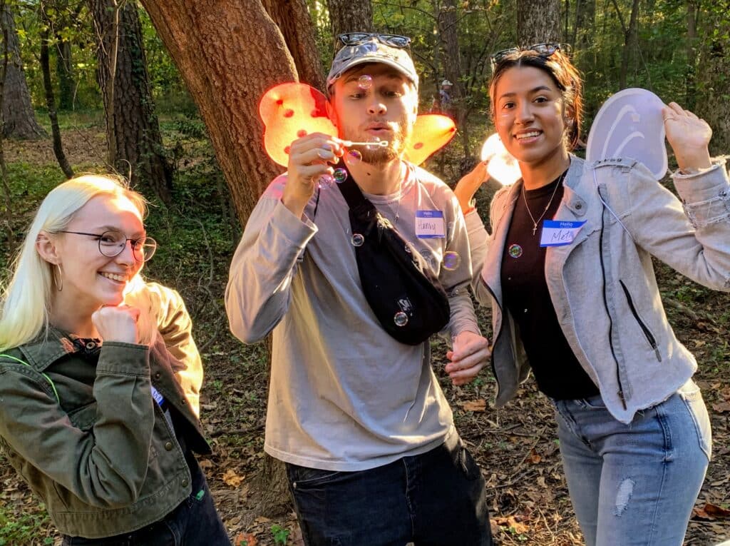 Three garden/nature club volunteers in a wooded area, blowing bubbles as the sunlight shines through the leaves.