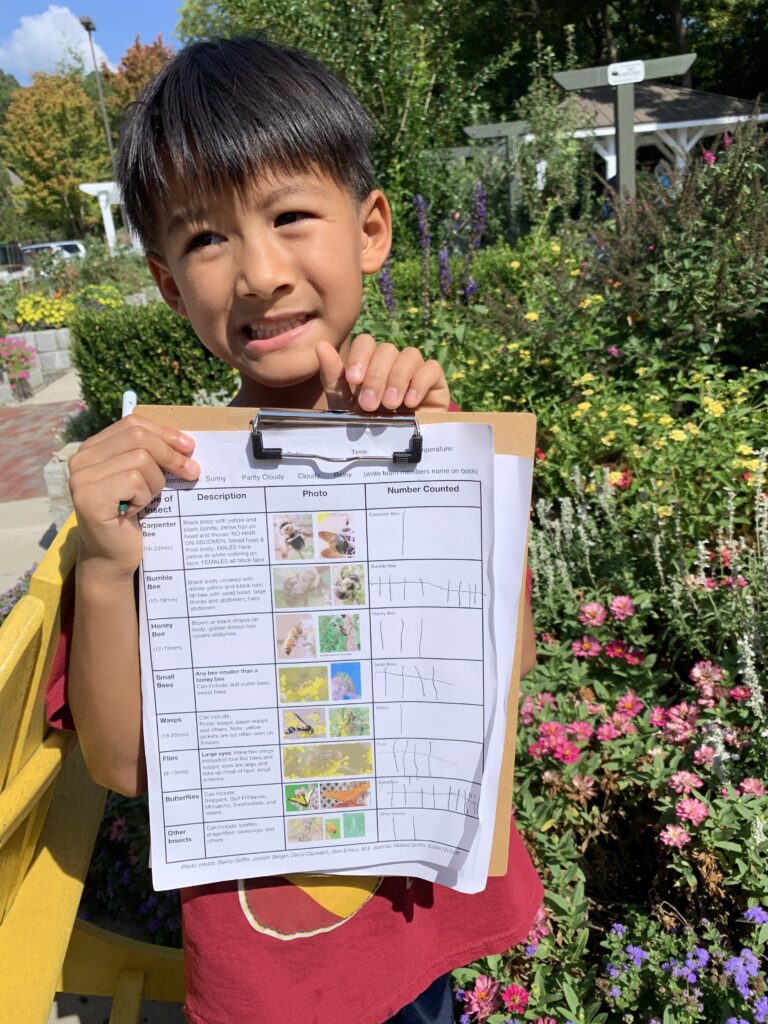 A young Asian boy standing in a garden holding up a worksheet attached to a clipboard, showing a garden class project he's doing.