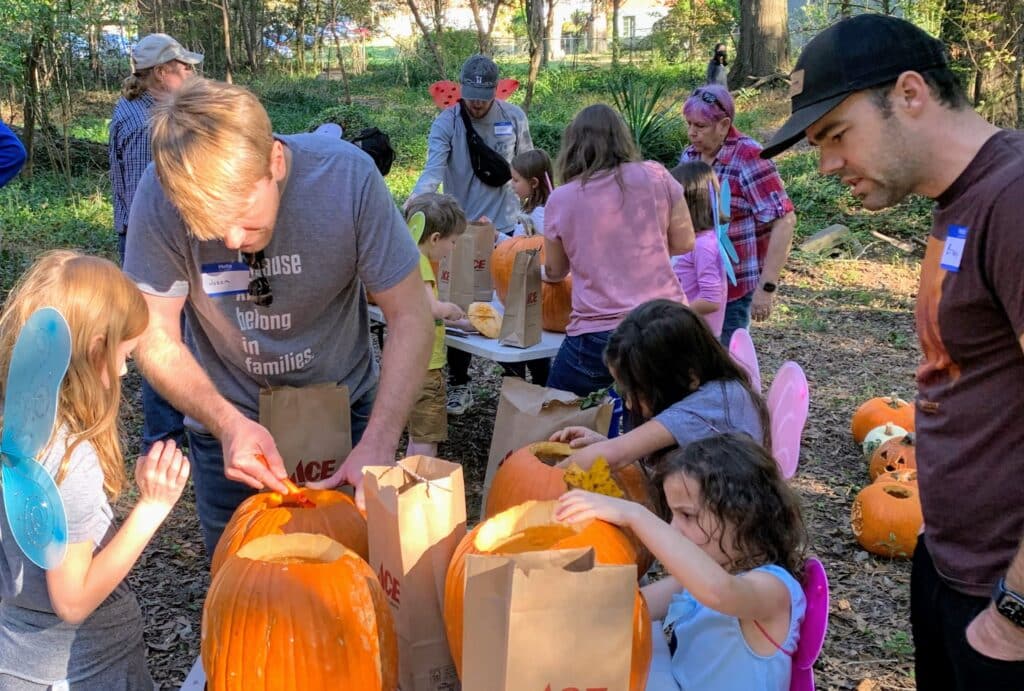 A group of kids with adult volunteers carving pumpkins in a wooded area.