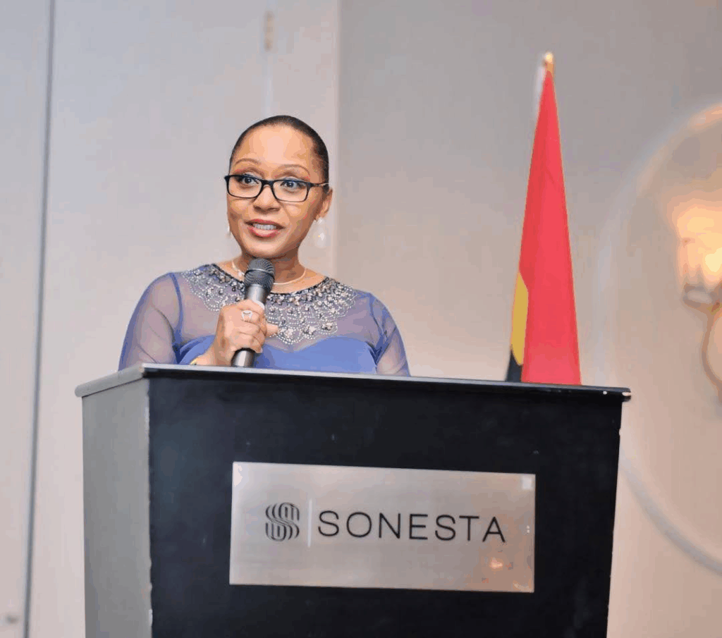 A young Black woman with her hair pulled back, wearing glasses and a purple dress. She's standing at a podium, holding a mic and speaking to the audience.