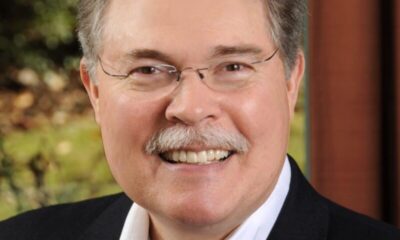 Mayor Mike Mason, wearing a dark jacket and white button-up shirt and glasses. He's standing in front of a window with lots of green trees and nature behind him.