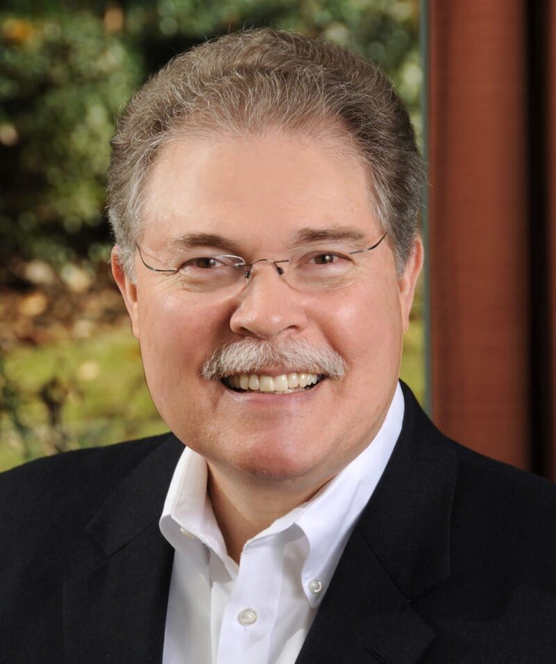 Mayor Mike Mason, wearing a dark jacket and white button-up shirt and glasses. He's standing in front of a window with lots of green trees and nature behind him.