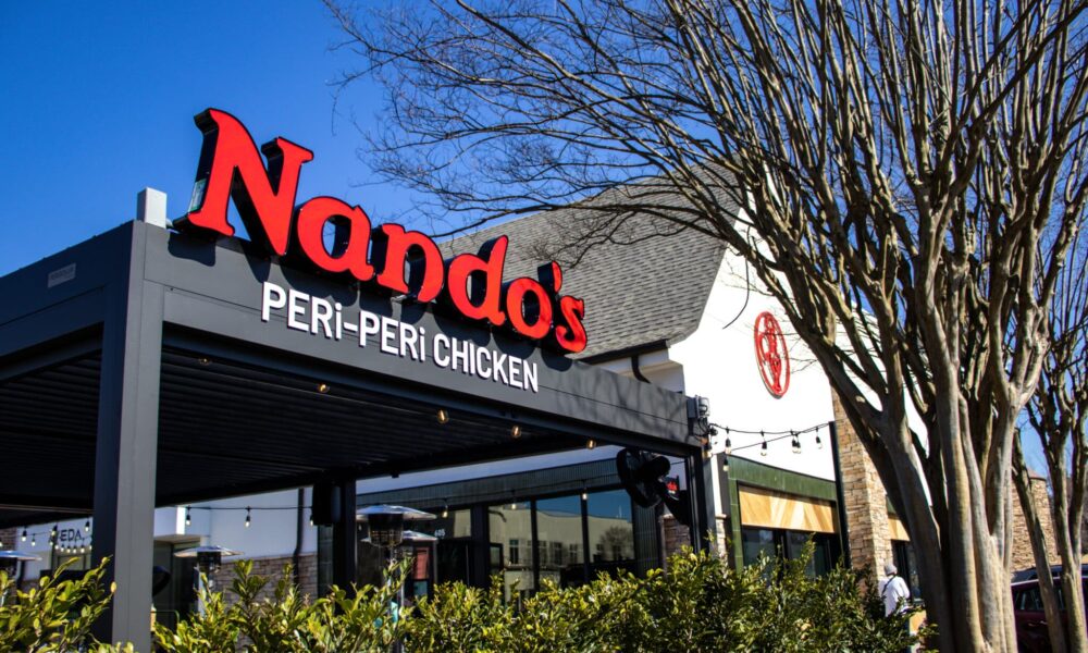 Exterior of a Nando's PERi-PERi chicken restaurant with signage, shrubs, tree and blue sky.