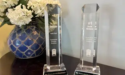 Two glass awards for best school headmaster and best workplace. They are on a dark wood table next to a blue vase with white flowers.