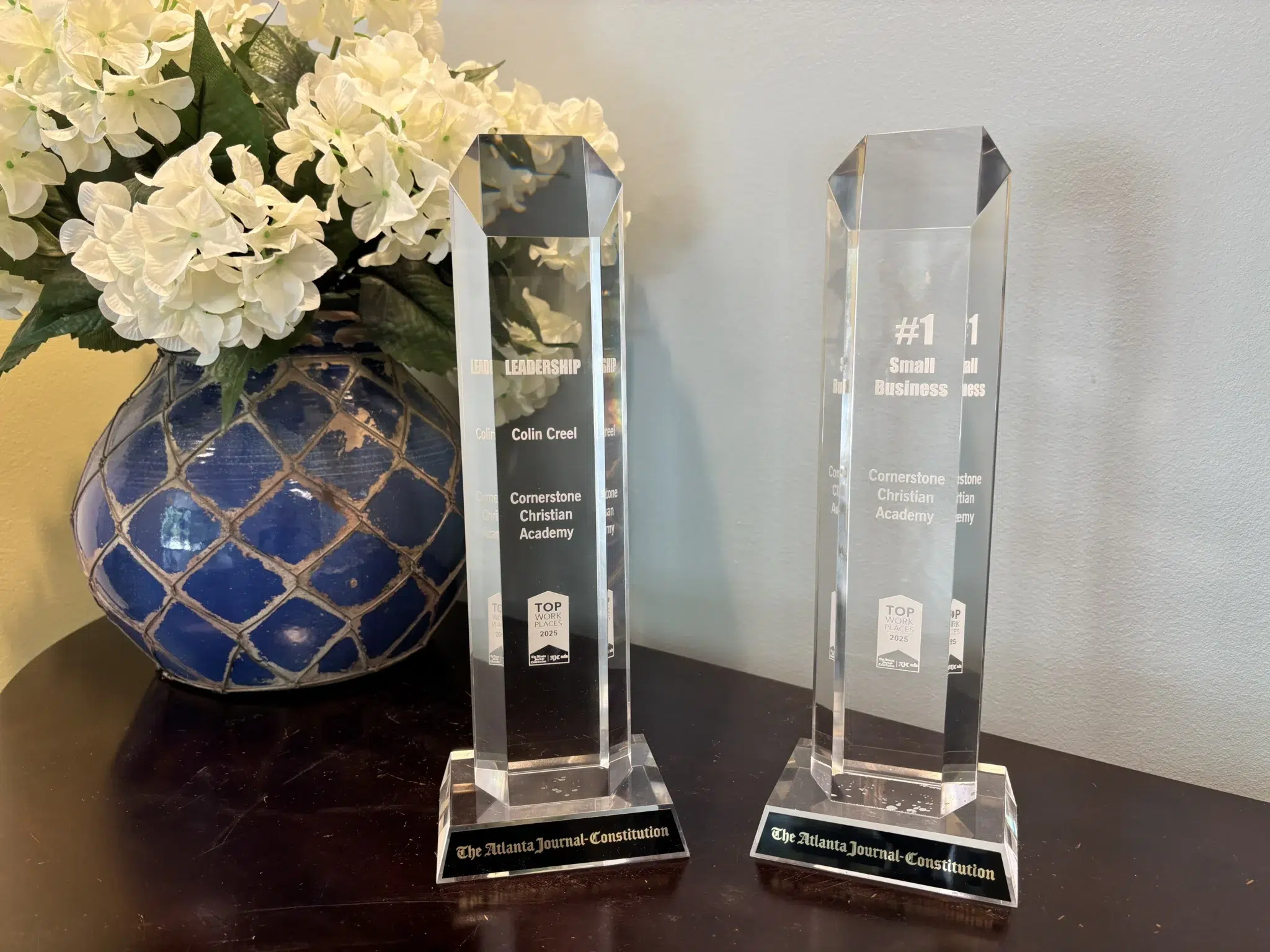 Two glass awards for best school headmaster and best workplace. They are on a dark wood table next to a blue vase with white flowers.