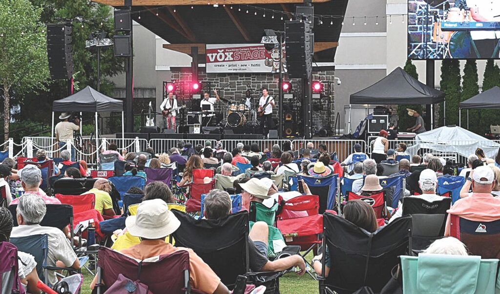 A large crowd of people sitting in lawn chairs watching a musical act perform on stage at an outdoor concert.