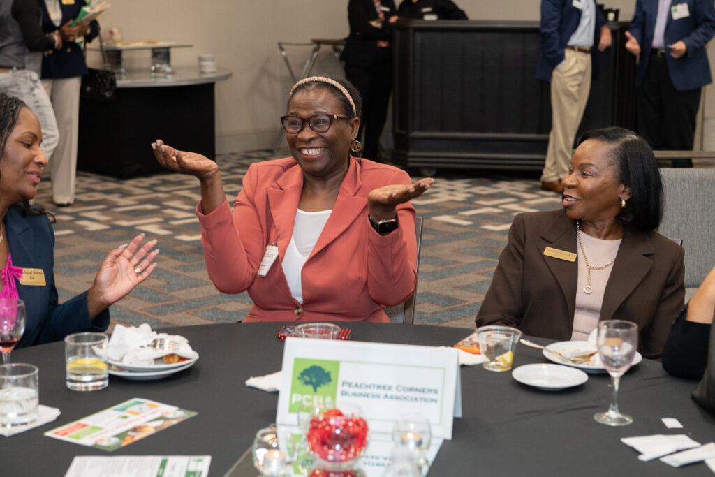 Three Black women are sitting at a table during a business event in a Peachtree Corners hotel conference room. They are smiling and the woman in the middle is gesturing with a shrug with her palms facing up.