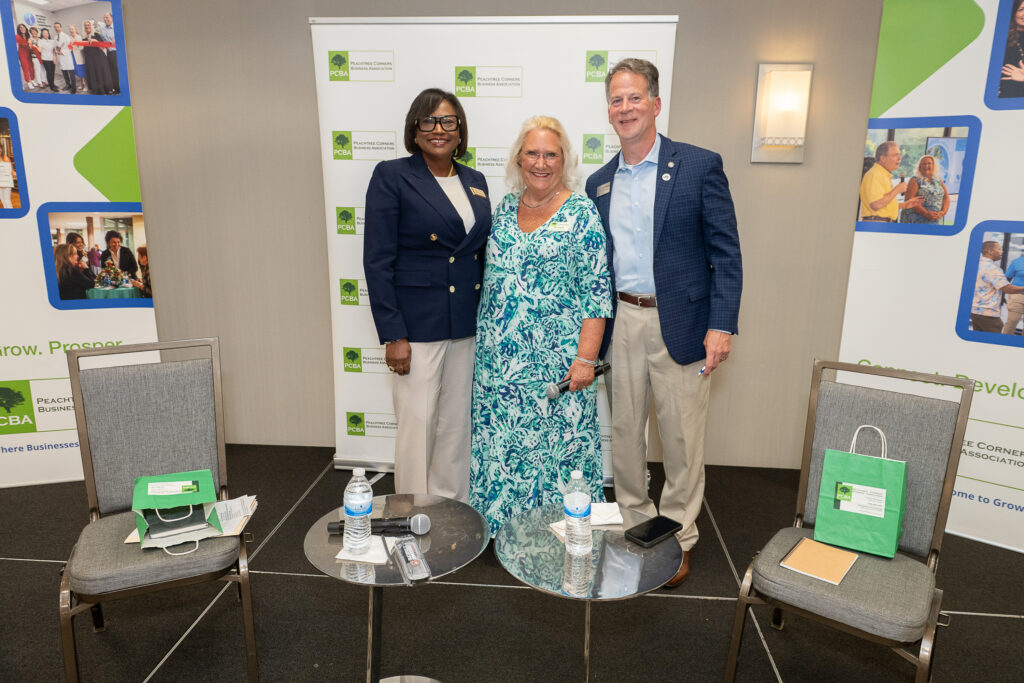 A Black woman, an older white woman with blonde hair and a white man in a blue blazer and khaki pants are standing together at a business event in a hotel conference room, posing for a photo.